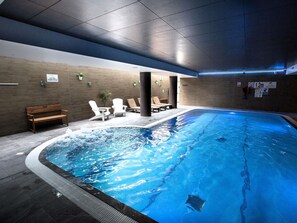 Une piscine intérieure dotée d’un banc et de chaises longues, entourée de murs ornés de motifs de carreaux et d’un banc en bois.