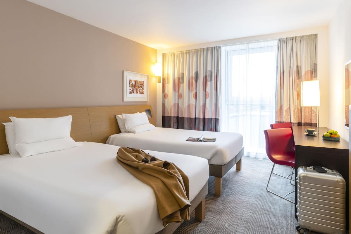 Habitación de hotel con dos camas, un escritorio, una silla, una lámpara y una maleta.