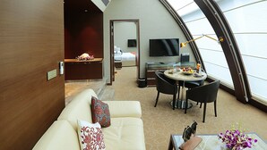 A modern hotel room with a curved ceiling, a sofa with decorative pillows, a dining area with a table and chairs, a flat-screen TV, and a large window providing natural light. - Sahara Star (Mumbai)
