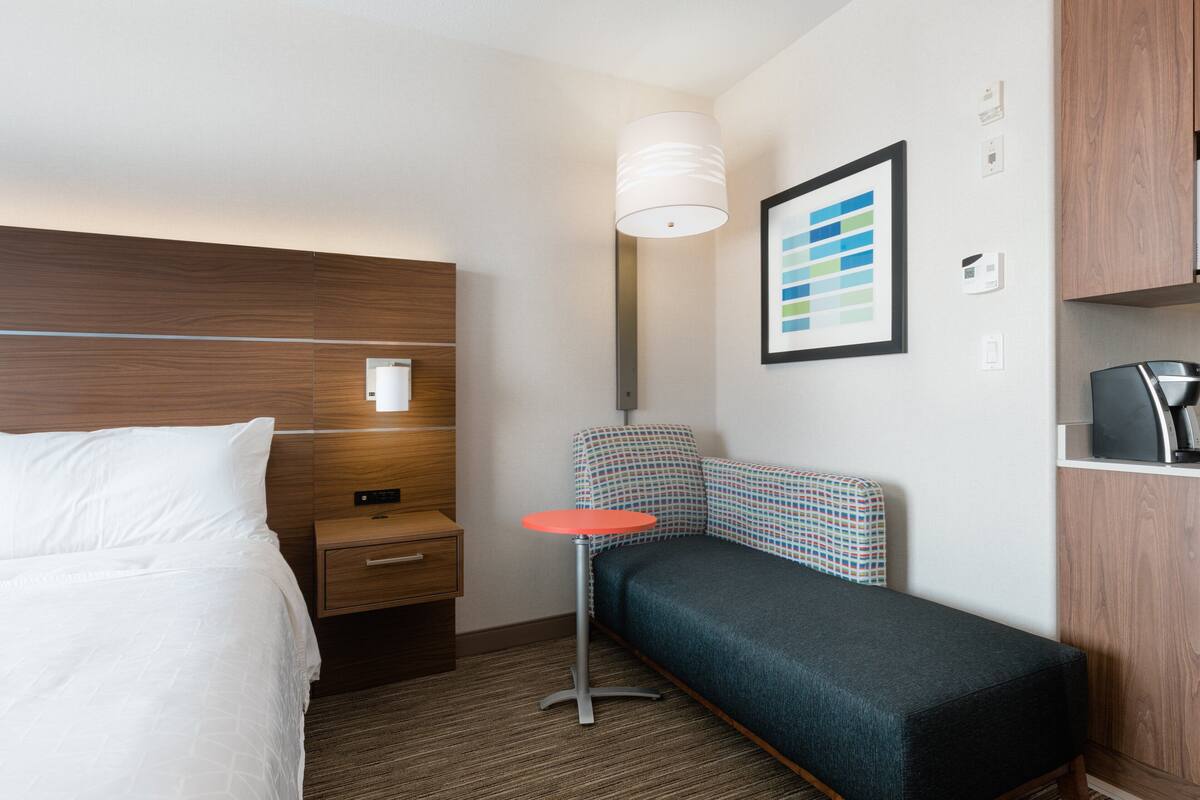 A hotel room with a bed, a bedside table, a small round table, a wall-mounted lamp, a colorful headboard, and a framed picture on the wall.