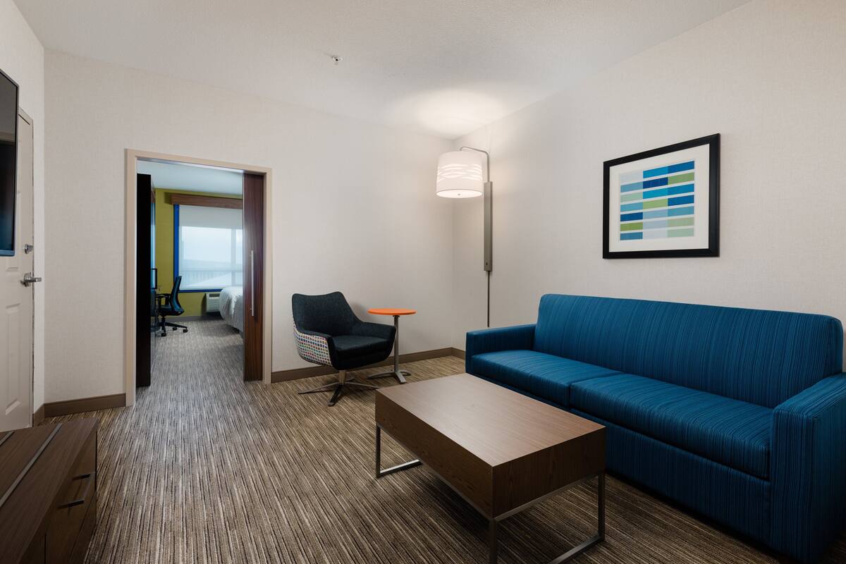 A hotel room with a blue sofa, a wooden coffee table, a black chair, a small orange table, and a framed abstract art piece on the wall.