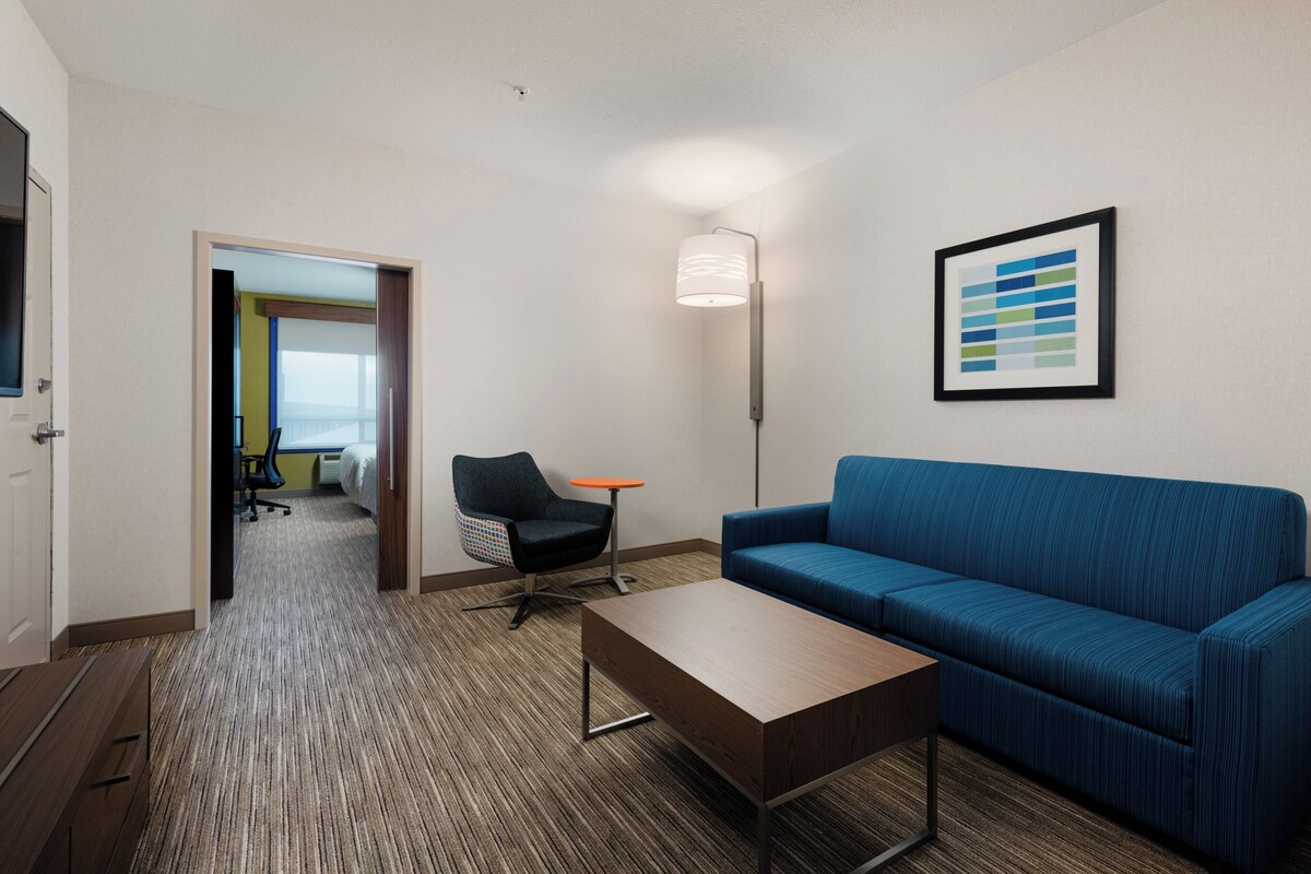 Habitación de hotel con un sofá azul, una mesa de centro de madera, una silla negra, una pequeña mesa naranja y una obra de arte abstracta enmarcada en la pared.