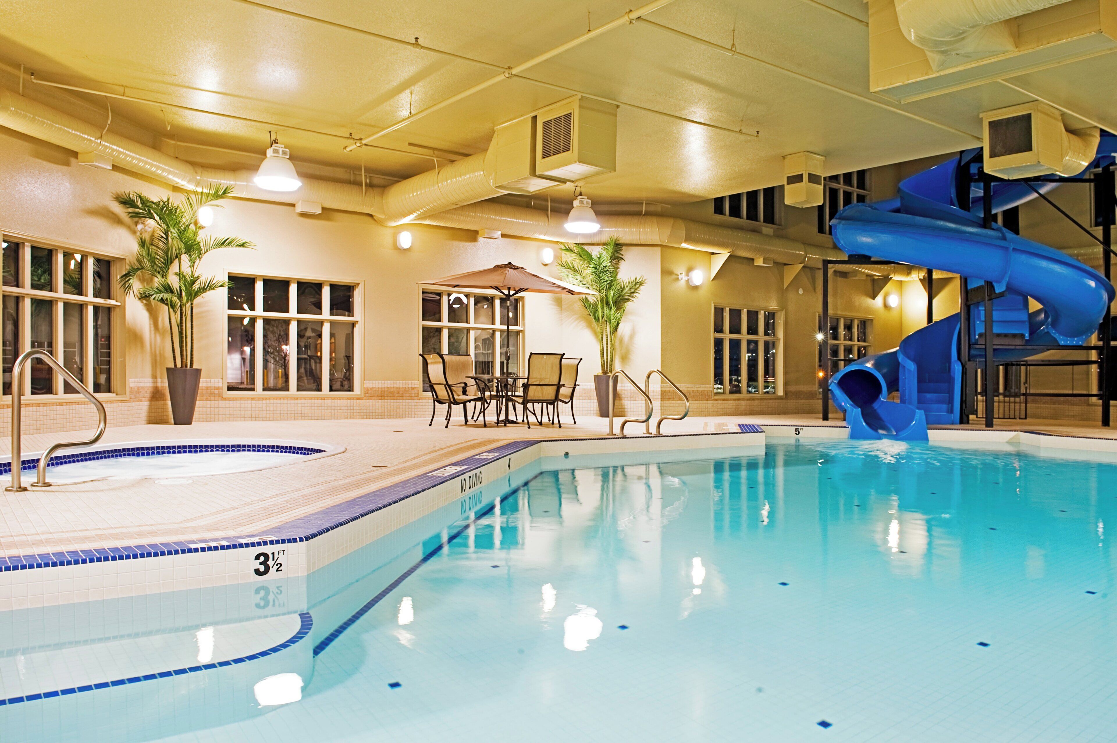 An indoor swimming pool with a blue slide, lounge chairs, and potted plants.