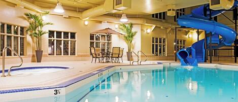 An indoor swimming pool with a blue slide, lounge chairs, and potted plants.