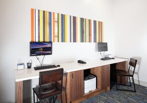 A hotel office space with two desks, each with a computer monitor, keyboard, and mouse. There are two chairs, a printer, and a small trash bin.