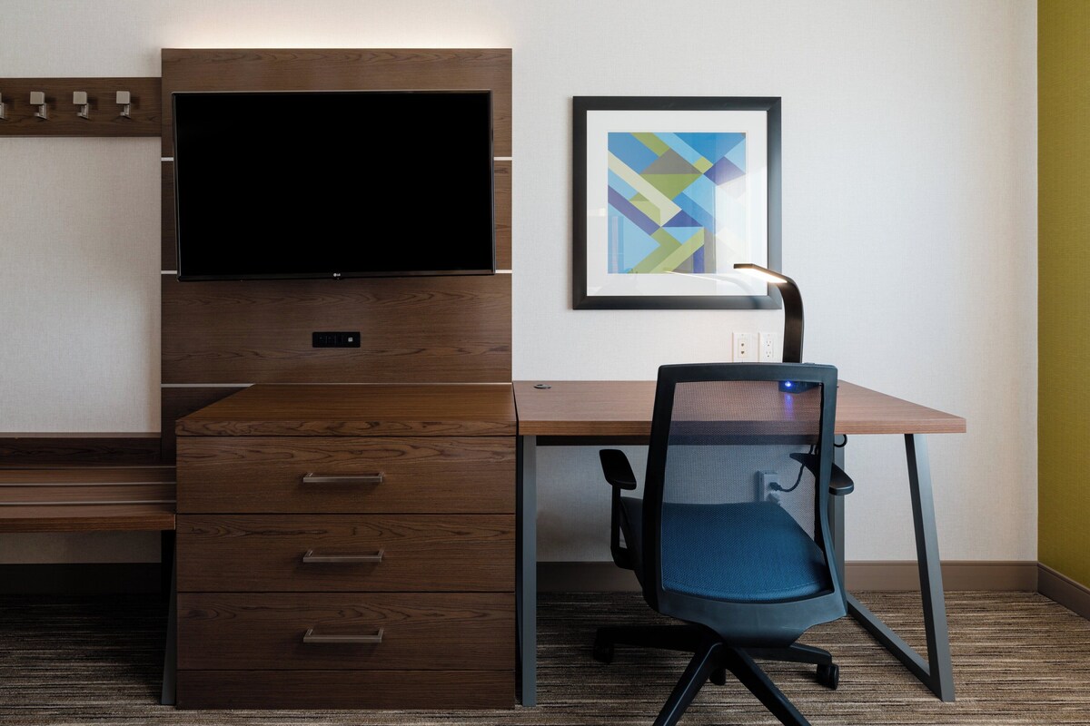 Una habitación de hotel con un escritorio de madera, un televisor, un cuadro abstracto enmarcado y una silla de oficina azul.