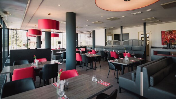 Een modern restaurantinterieur met hoge plafonds, rode en zwarte stoelen en gedekte tafels.