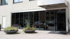 Een modern gebouw met een glazen entree, een bord met de tekst 'BASEION HOTEL' en twee grote plantenbakken met bloemen ervoor.