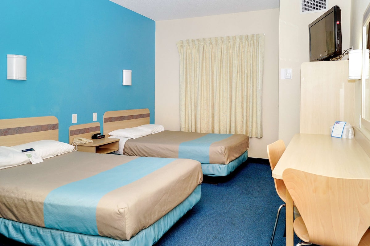 Ein Hotelzimmer mit zwei Betten, einem Schreibtisch, einem Fernseher und einem Fenster mit Vorhängen.