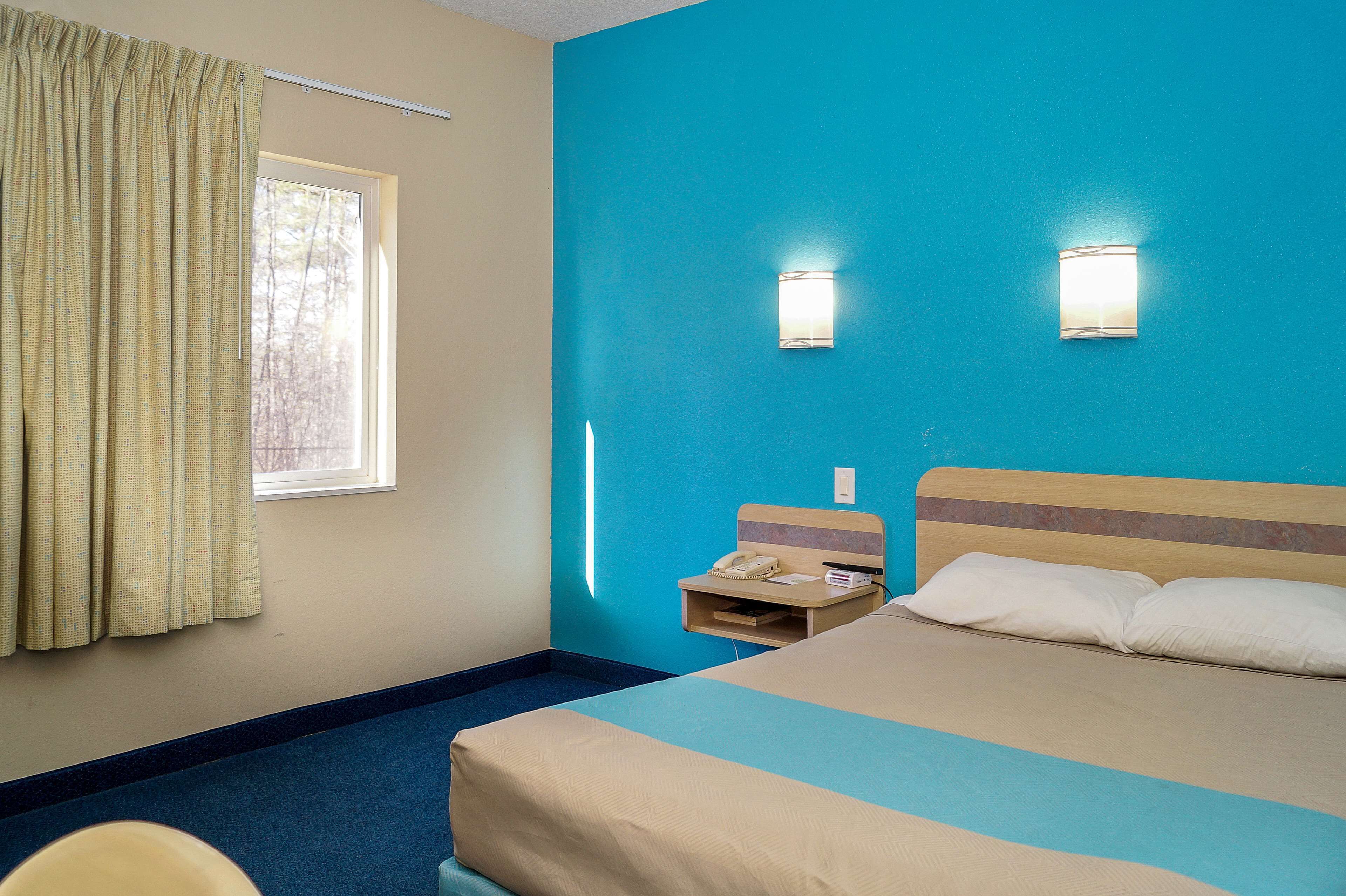 Habitación de hotel con cama, mesita de noche, ventana con cortinas y dos luces en la pared.