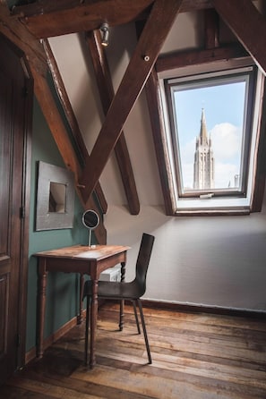 Una habitación con techo inclinado, vigas de madera, un escritorio de madera, una silla y una ventana con vista a un edificio.