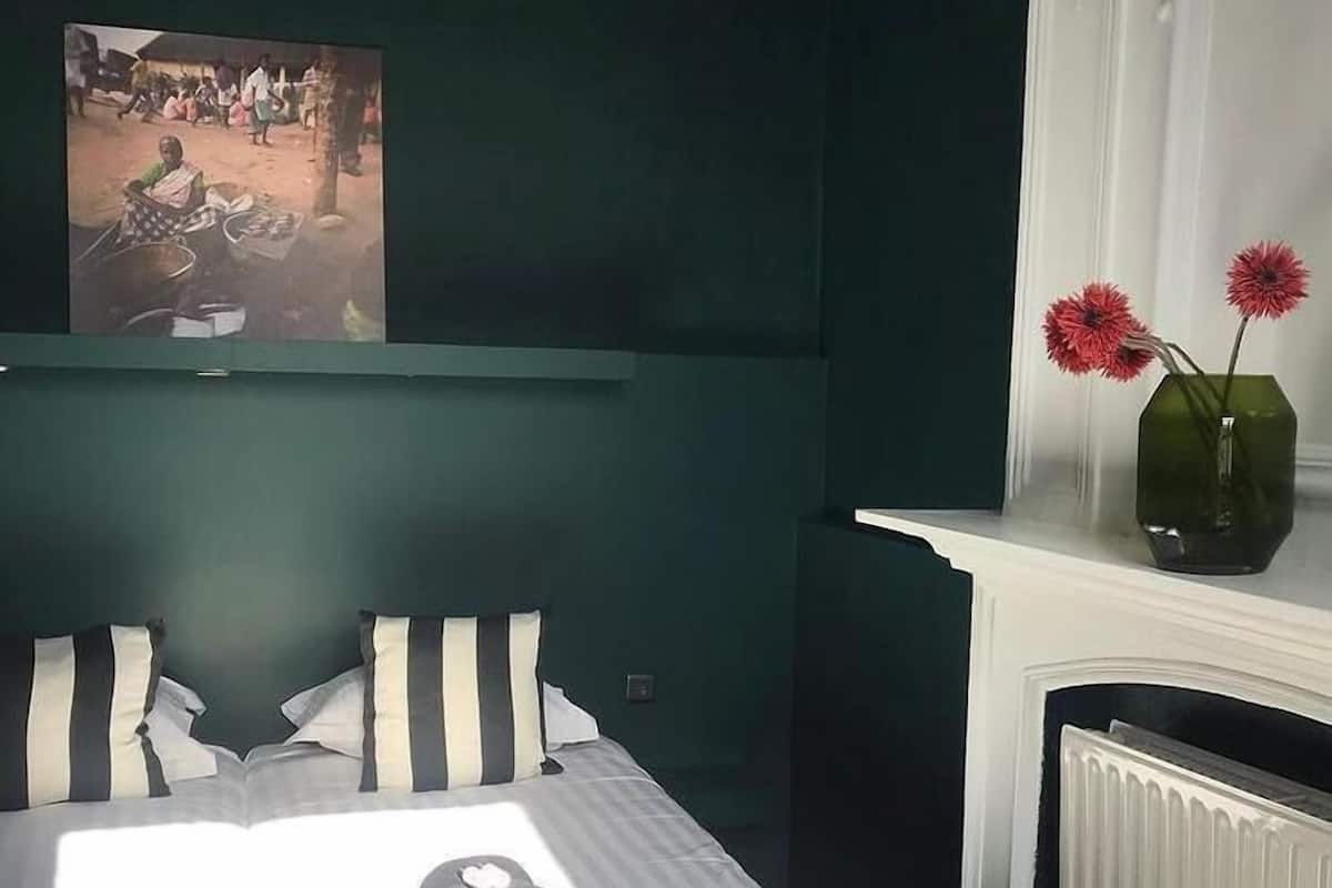 Une chambre à coucher avec un lit, des oreillers, un livre, un vase de fleurs, une cheminée et un tableau au mur.