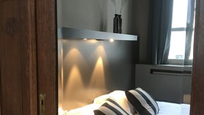 Een slaapkamer met een bed, kussens, een nachtkastje, een raam met gordijnen, en een wandplank met twee flessen.