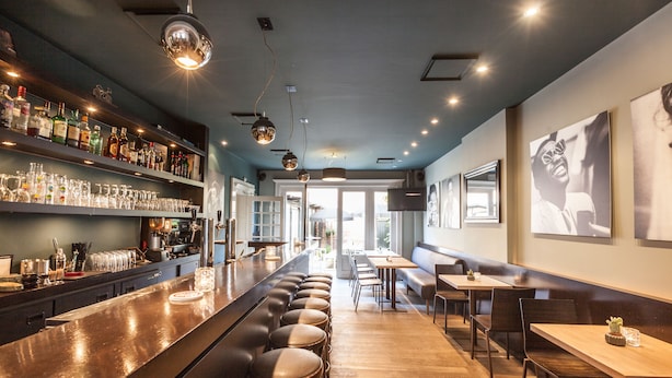 Een bar met een lange toonbank, krukken en flessenplanken. Achtergrond bevat tafels en stoelen, en een groot raam is zichtbaar.