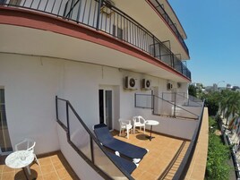 Een balkon met een blauwe bank, witte stoelen en een kleine tafel. Er staan airconditioners aan de muur en er is uitzicht op de stad.