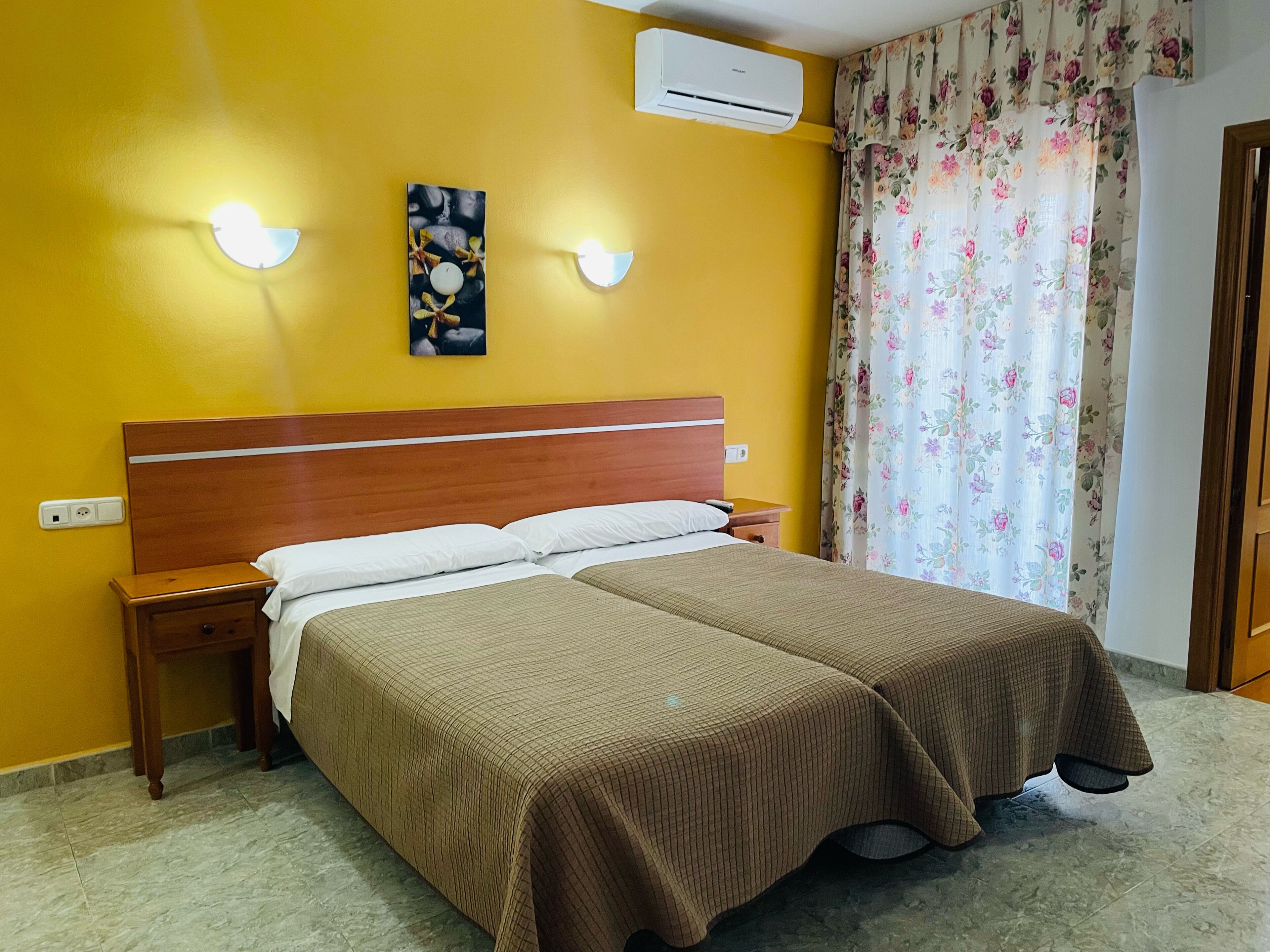 Ett hotellrum med en sänggavel i trä, två sängar med bruna sängkläder, ett nattduksbord, en luftkonditioneringsanläggning och en blommönstrad gardin.