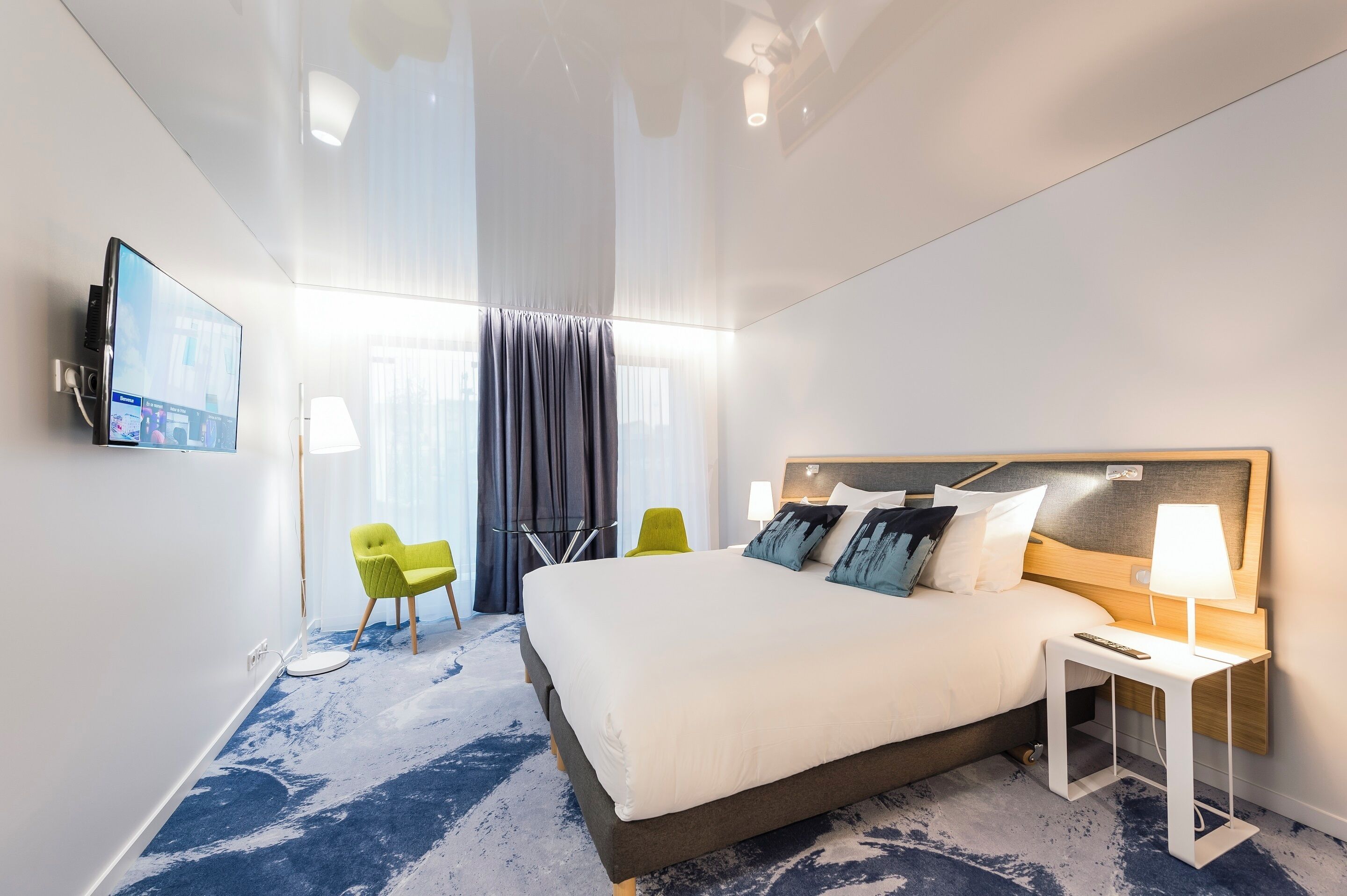 A hotel room with a large bed, a TV mounted on the wall, a desk with a lamp, and a chair.