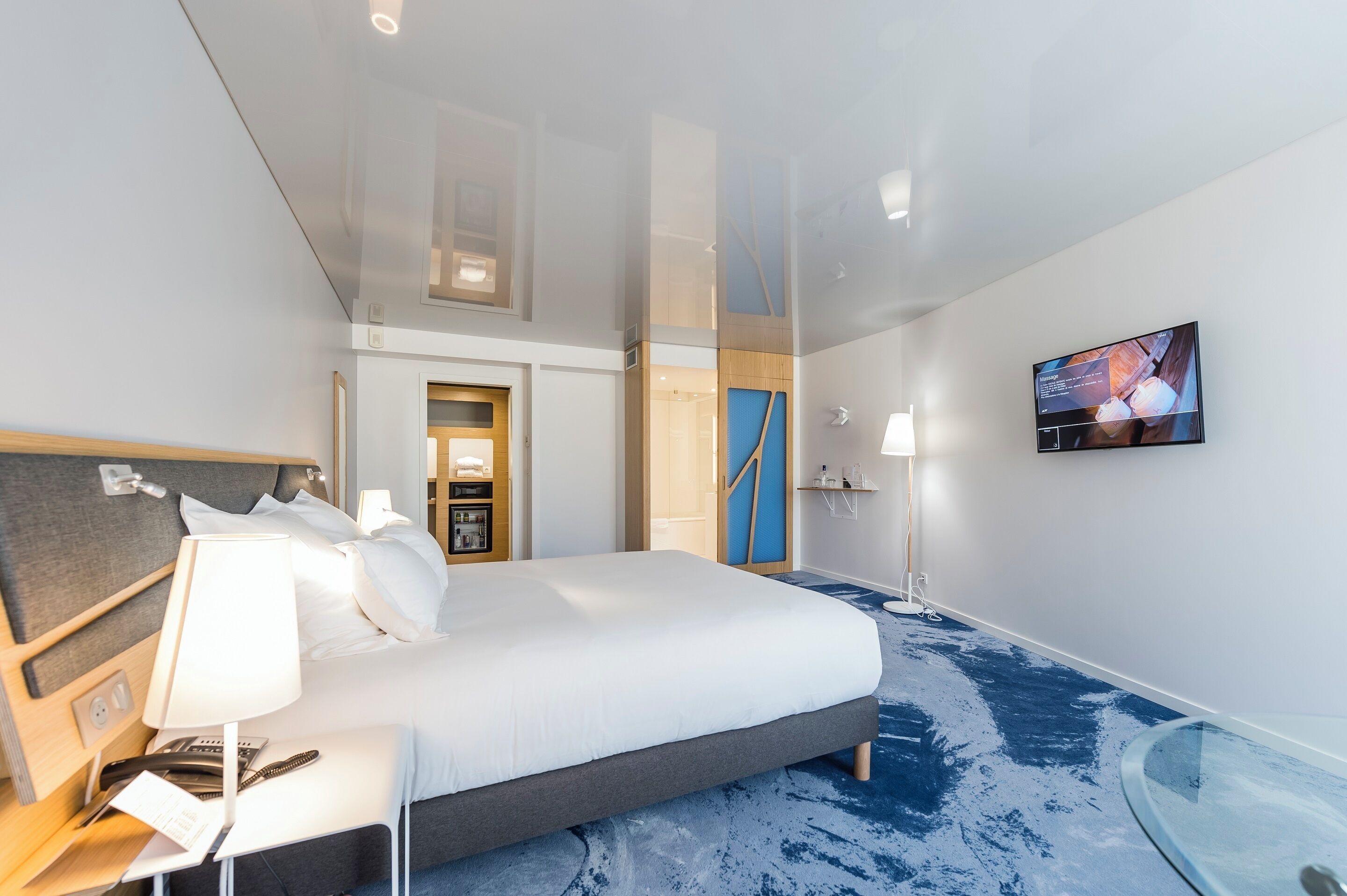 A modern hotel room with a large bed, bedside table with lamp, and a TV on the wall.