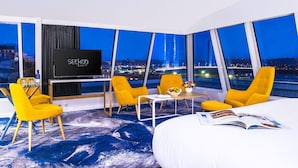 Une chambre d’hôtel moderne avec un grand lit, une télévision, deux fauteuils jaunes, une petite table sur laquelle se trouve un saladier rempli de fruits, et une vue sur le paysage urbain illuminé la nuit.