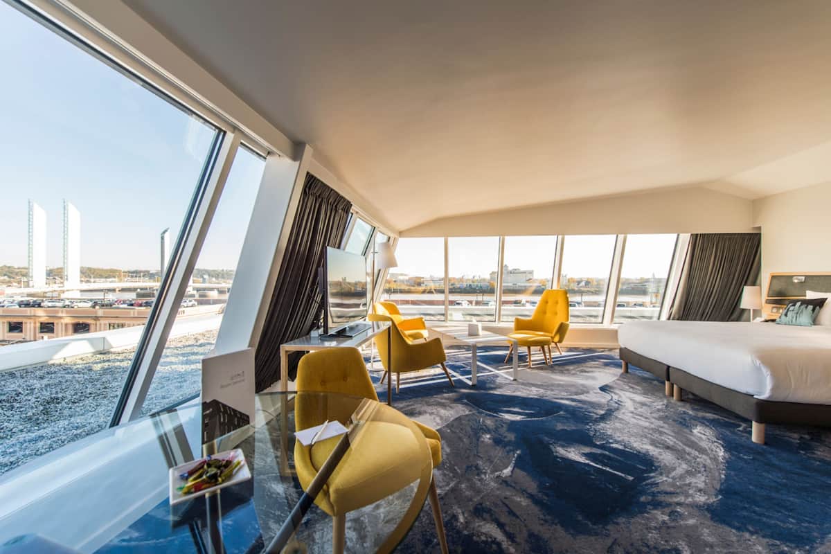 Una habitación de hotel con un ventanal grande, una cama, un escritorio y dos sillas.