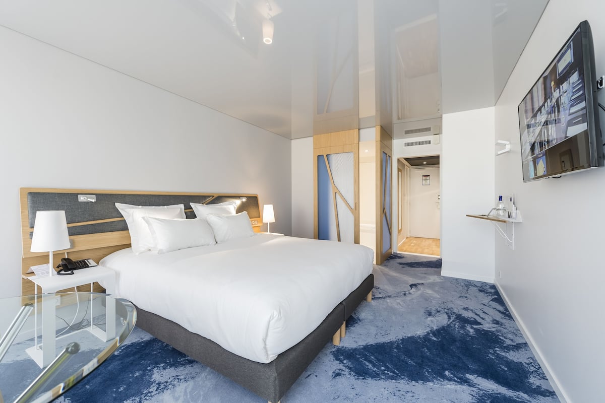 Una habitación de hotel moderna con una cama grande, una mesita de vidrio, un televisor montado en la pared y una alfombra azul.