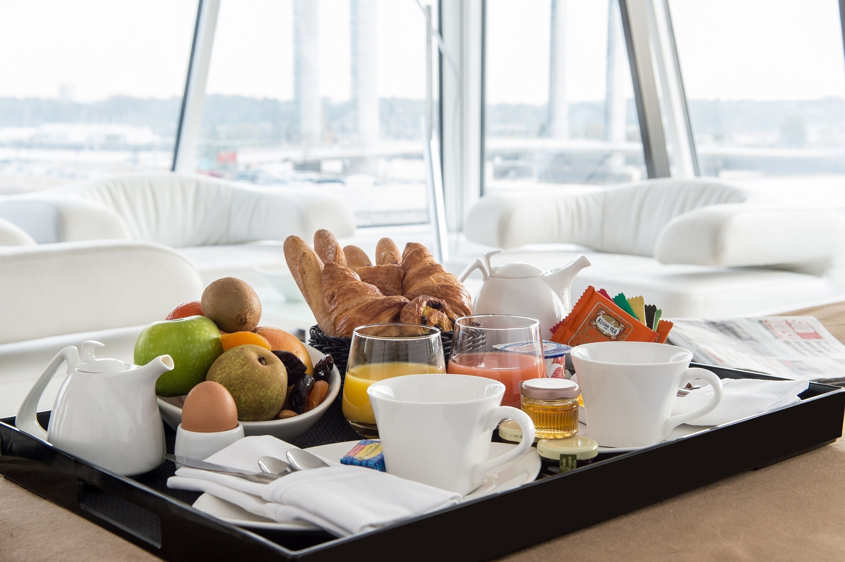 A breakfast tray with croissants, fruits, eggs, juice, coffee, and tea on a hotel room table with white furniture and a city view.