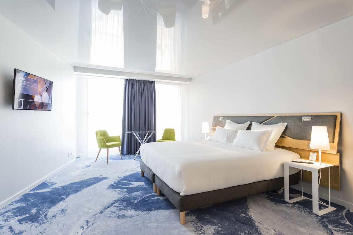Una habitación de hotel con una cama grande, una mesita de noche con lámpara, un televisor en la pared y una ventana con cortinas.