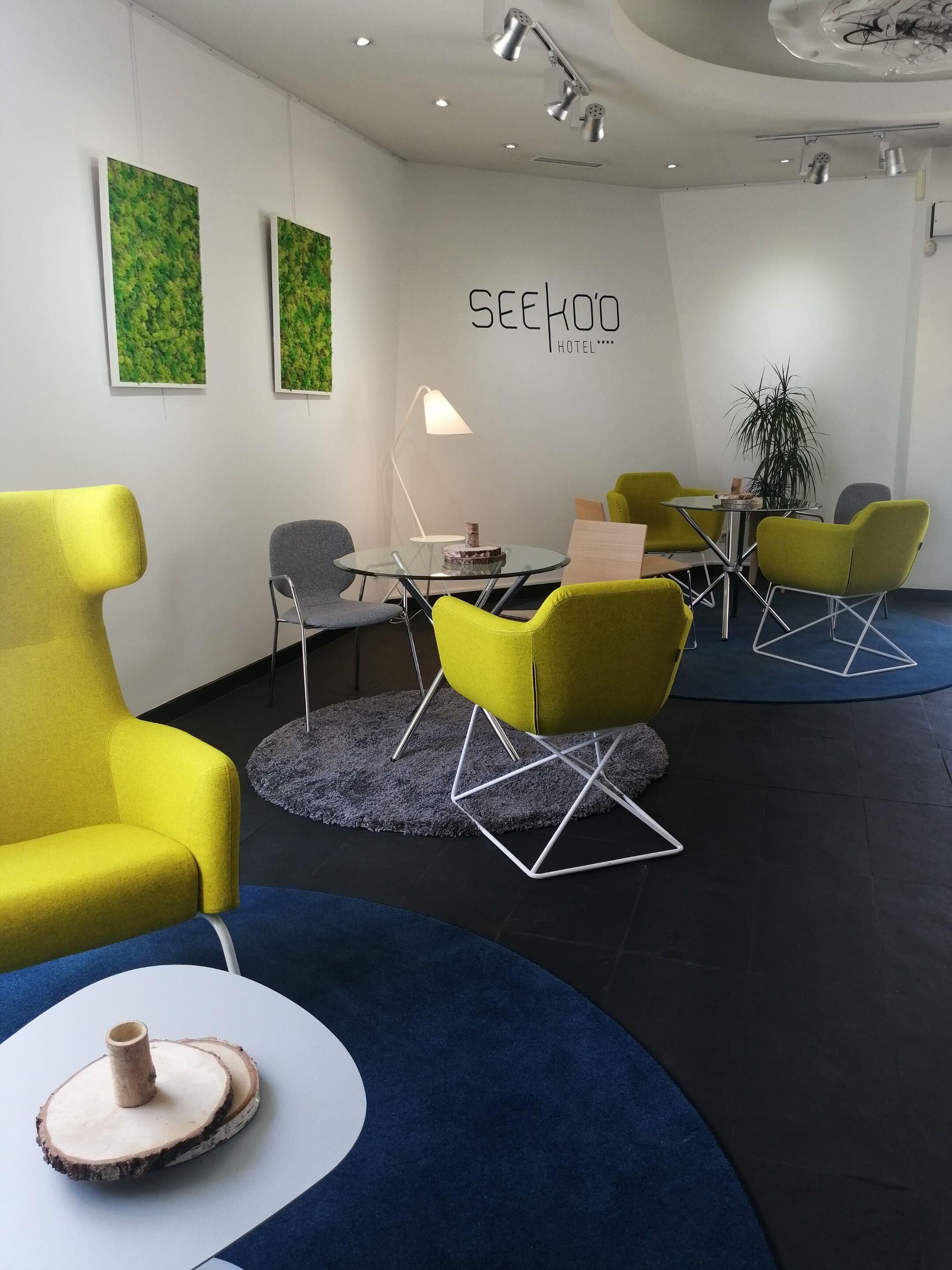 Una habitación de hotel moderna con una mesa redonda de vidrio, sillas amarillas y verdes, una pequeña mesa redonda con un florero y un cartel en la pared.
