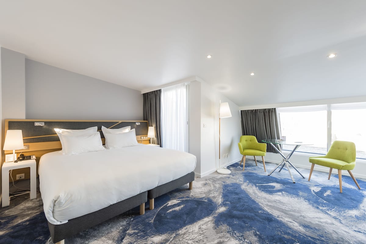 Una habitación de hotel moderna con una cama grande, dos sillas, una mesita y una ventana con cortinas.