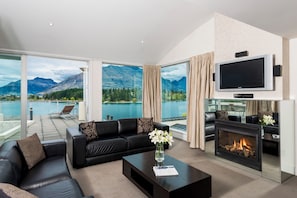A modern living room with a fireplace, a black leather sofa, a coffee table with a vase of flowers, and a large TV mounted on the wall. - Oaks Queenstown Club Suites (Queenstown)