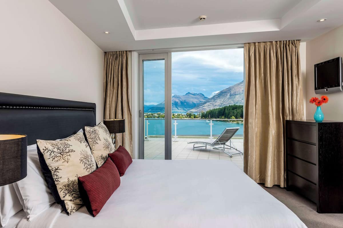 Ett hotellrum med en stor säng, en tv, en byrå och utsikt över en sjö och berg.