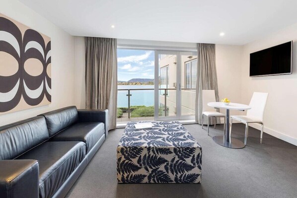 A modern hotel room with a black leather sofa, a patterned ottoman, a small round table with two chairs, a flat-screen TV, and a balcony with a view of the sea.