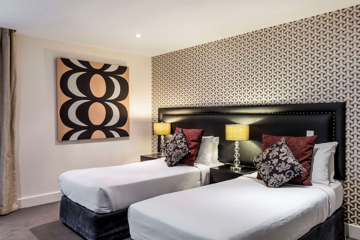 Ett hotellrum med två sängar, en stor, abstrakt målning, tapet med mönster och sänglampor.