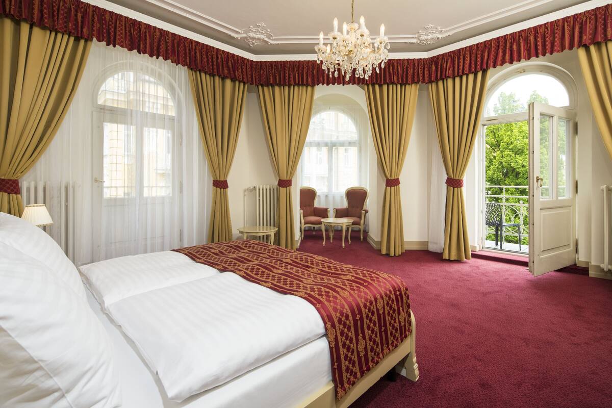 Une chambre d’hôtel avec un grand lit, un lustre, un coin salon avec des fauteuils et de grandes fenêtres avec des rideaux.