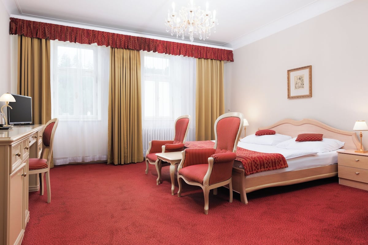 Une chambre d’hôtel avec un grand lit, deux fauteuils, un bureau avec une télévision et un lustre.