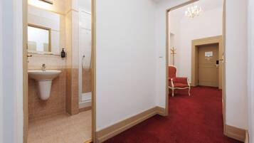 A hotel corridor with a bathroom on the left, a red carpeted area, and a door with a sign.
