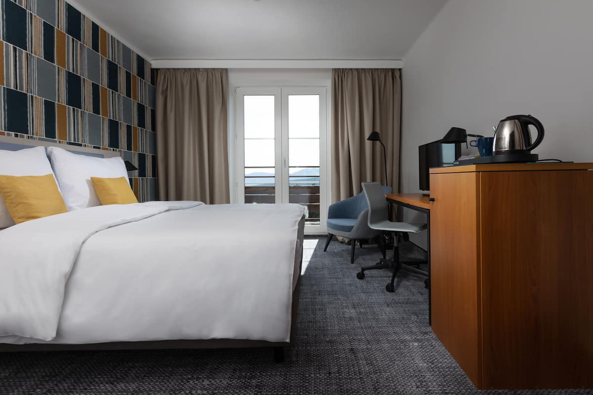 A hotel room with a bed, a desk with a laptop, a chair, a wardrobe, and a view of the sea through the window.