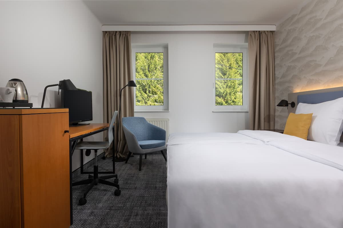 A hotel room with a bed, a desk with a computer, a chair, and two windows with curtains.