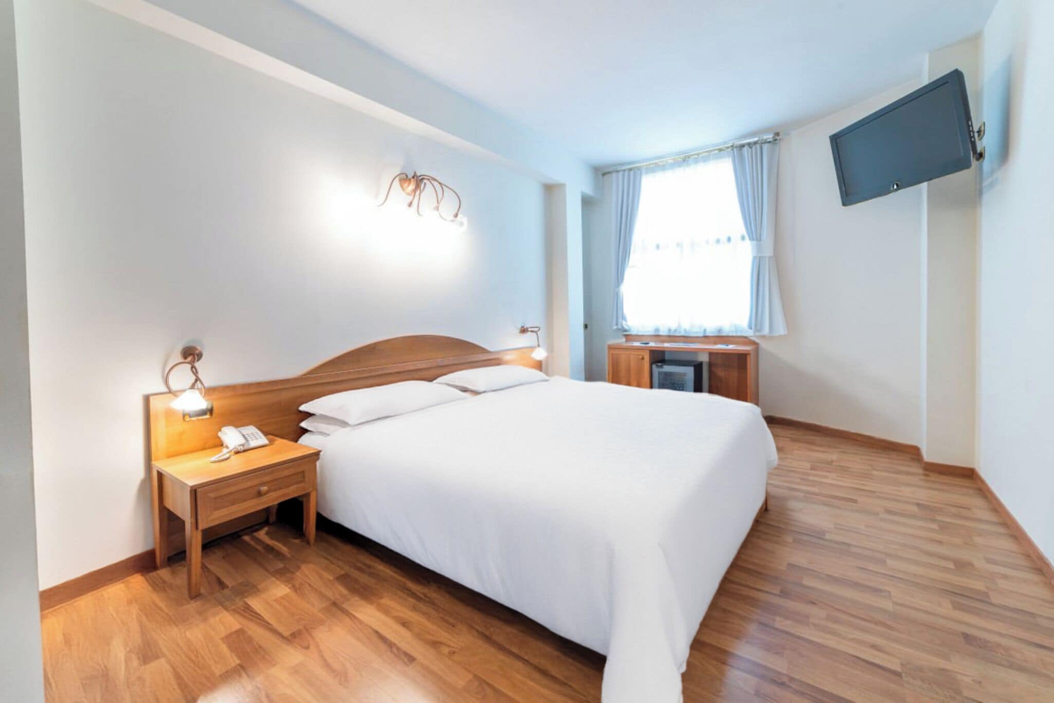 Une chambre d’hôtel avec un lit, une table de chevet, une lampe, une télévision et une fenêtre avec des rideaux.