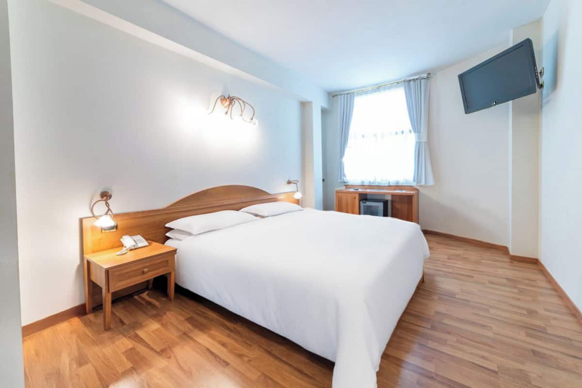 A hotel room with a bed, bedside table, lamp, TV, and a window with curtains.
