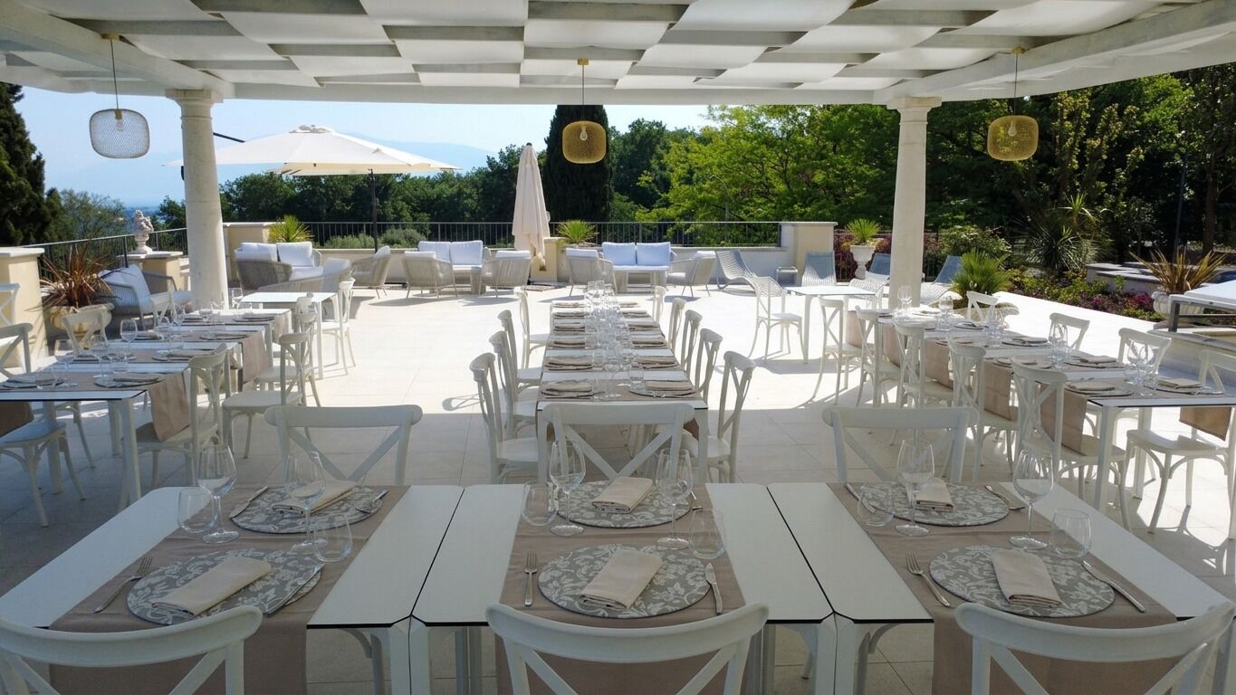 An outdoor dining area with white tables and chairs, arranged in rows, under a pergola with a view of greenery and a distant landscape.