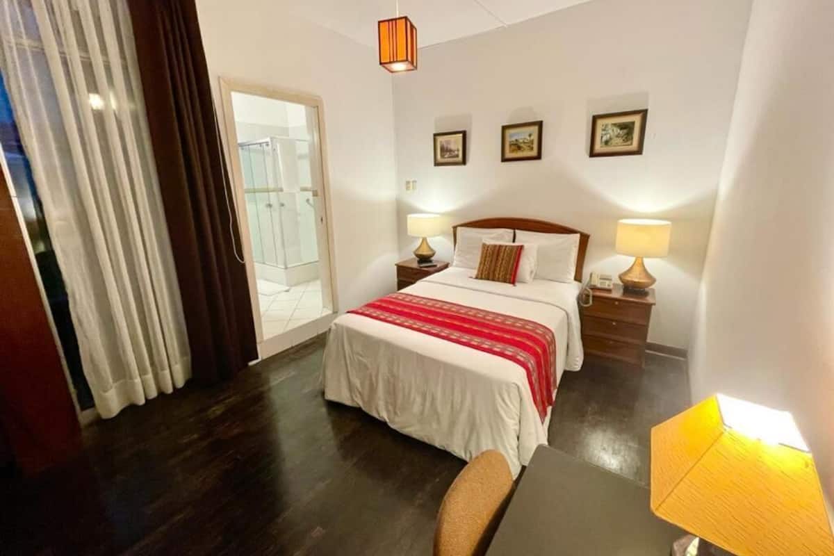 A hotel room with a bed, two bedside tables with lamps, a chair, and a view of the outside through a glass door.