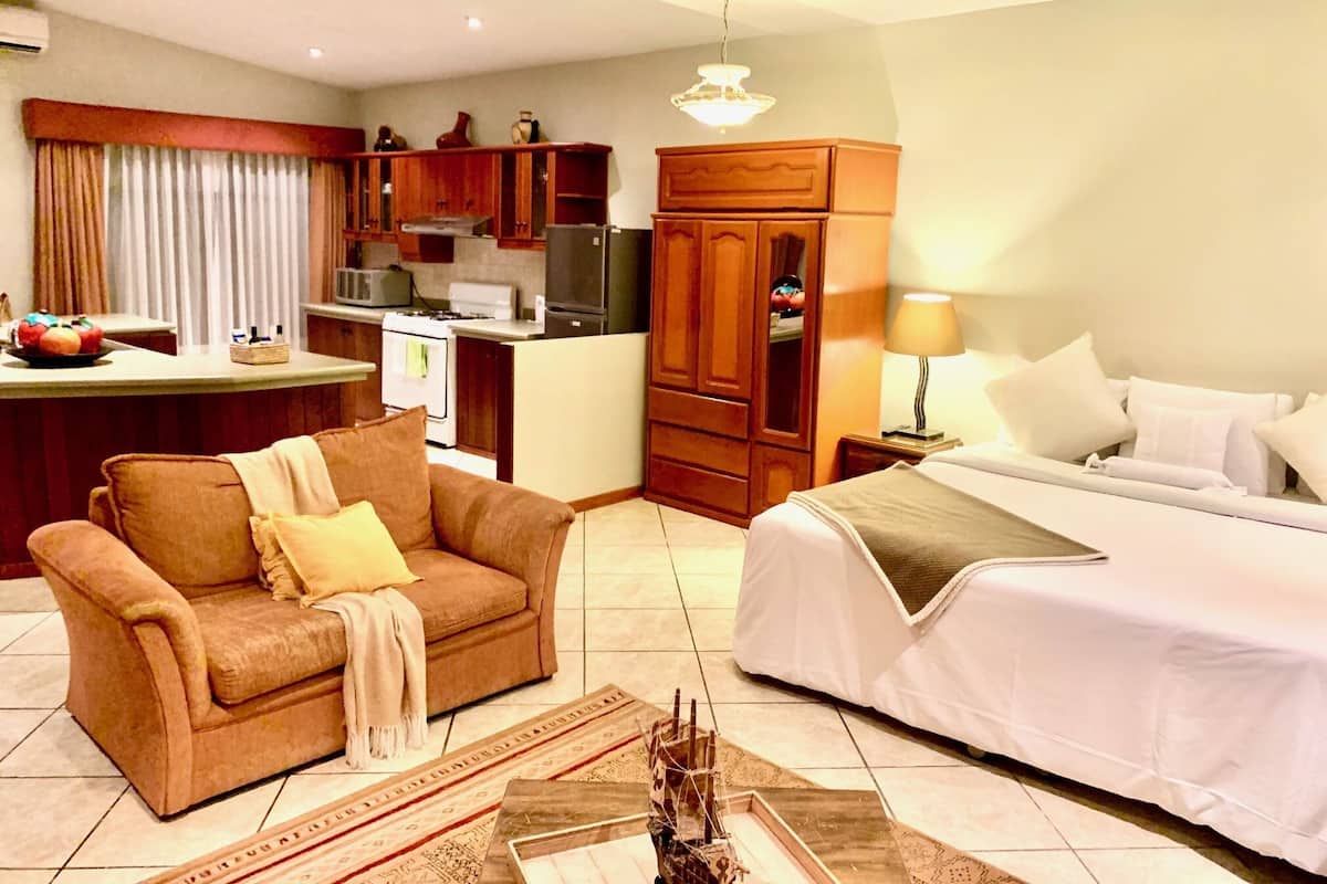 A compact hotel room with a kitchenette, a sofa, a bed, and a nightstand with a lamp.