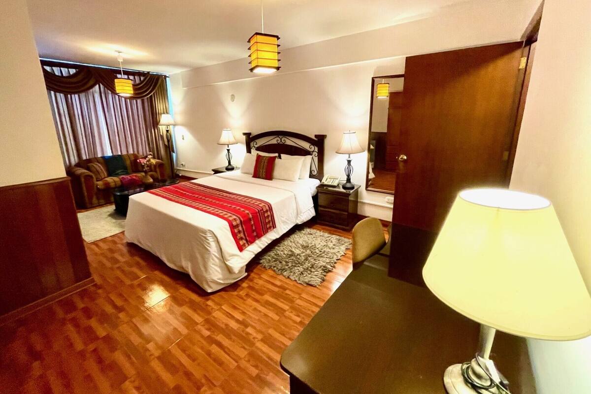 A hotel room with a neatly made bed, a desk with a lamp, a chair, a sofa, and a mirror.