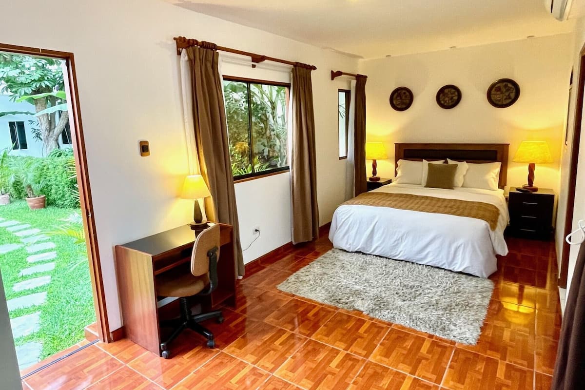 A hotel room with a bed, desk, and chair, a small rug, and a view of a garden.