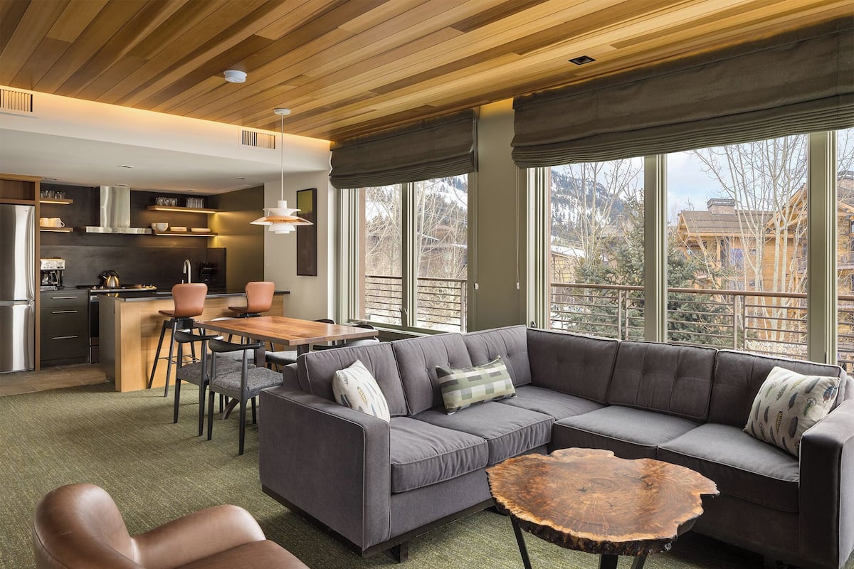 A modern kitchen with a central island, a dining area with a wooden table and chairs, a gray sofa with cushions, and large windows offering a view of the outside.