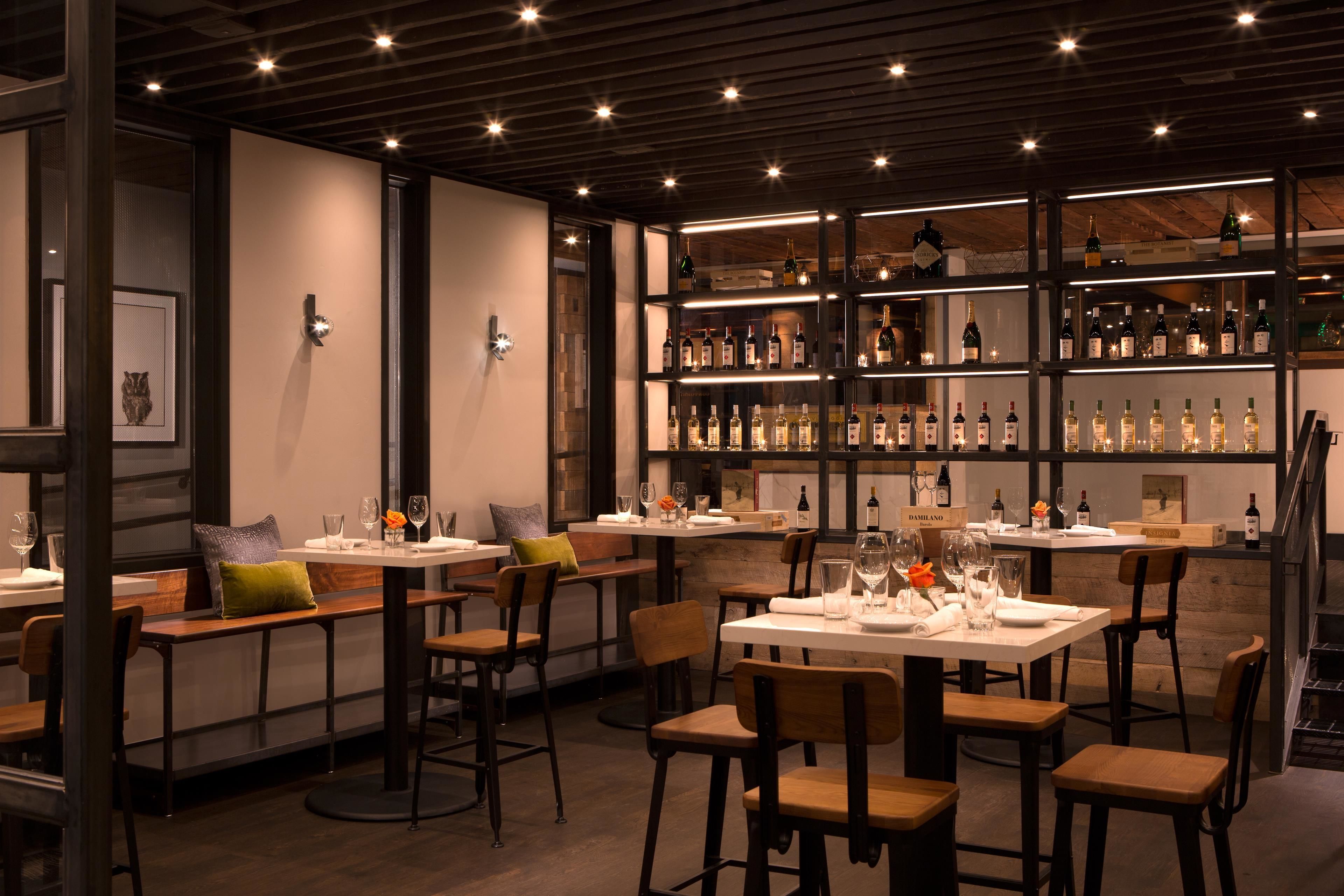 Un interior de restaurante con mesas y sillas de madera, una estantería con botellas de vino y obras de arte enmarcadas en las paredes.