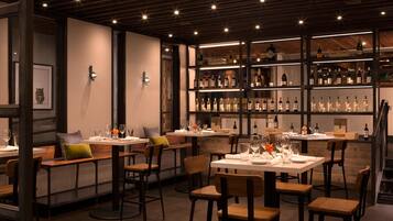 Un interior de restaurante con mesas y sillas de madera, una estantería con botellas de vino y obras de arte enmarcadas en las paredes.