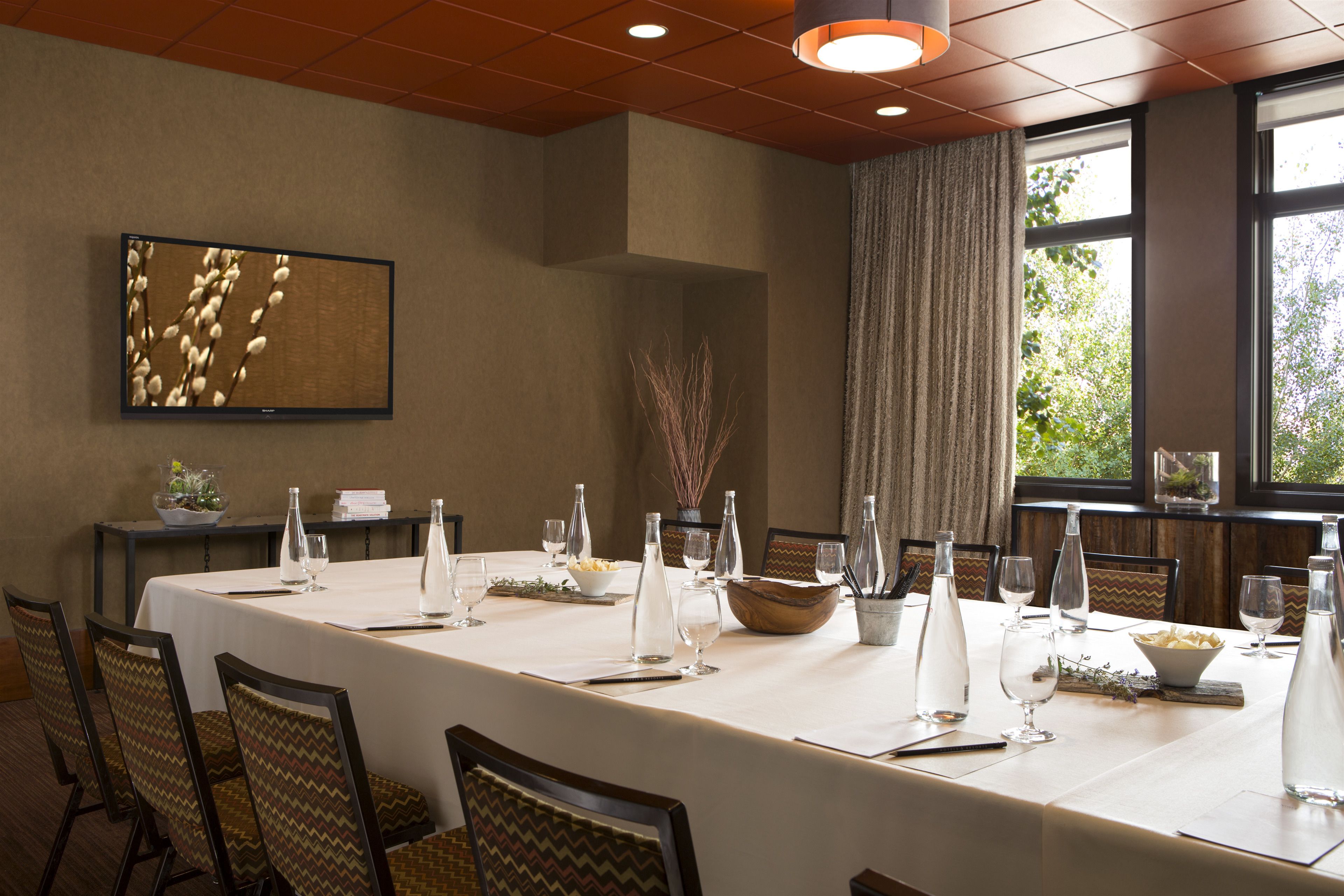 A conference room with a long table set for a meeting, chairs, a large window with a view, and a wall-mounted screen.
