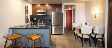 A modern kitchen with a central island, bar stools, a dining area with chairs, and a refrigerator.